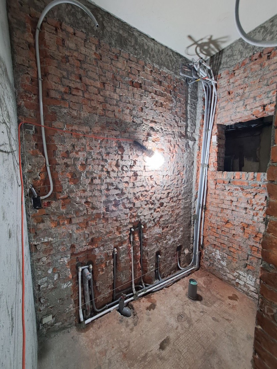 跨場域水電施工現場：老屋打除粉刷層後可見裸露磚牆，地坪沿牆腳重新埋設給排水配管，壁面與天花板可見新拉的電氣管路 rough-in 階段。
