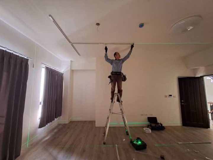 設配安裝:師傅站在鋁梯上於客廳天花板安裝燈具線槽,地面以雷射水平儀打出綠色基準線。
